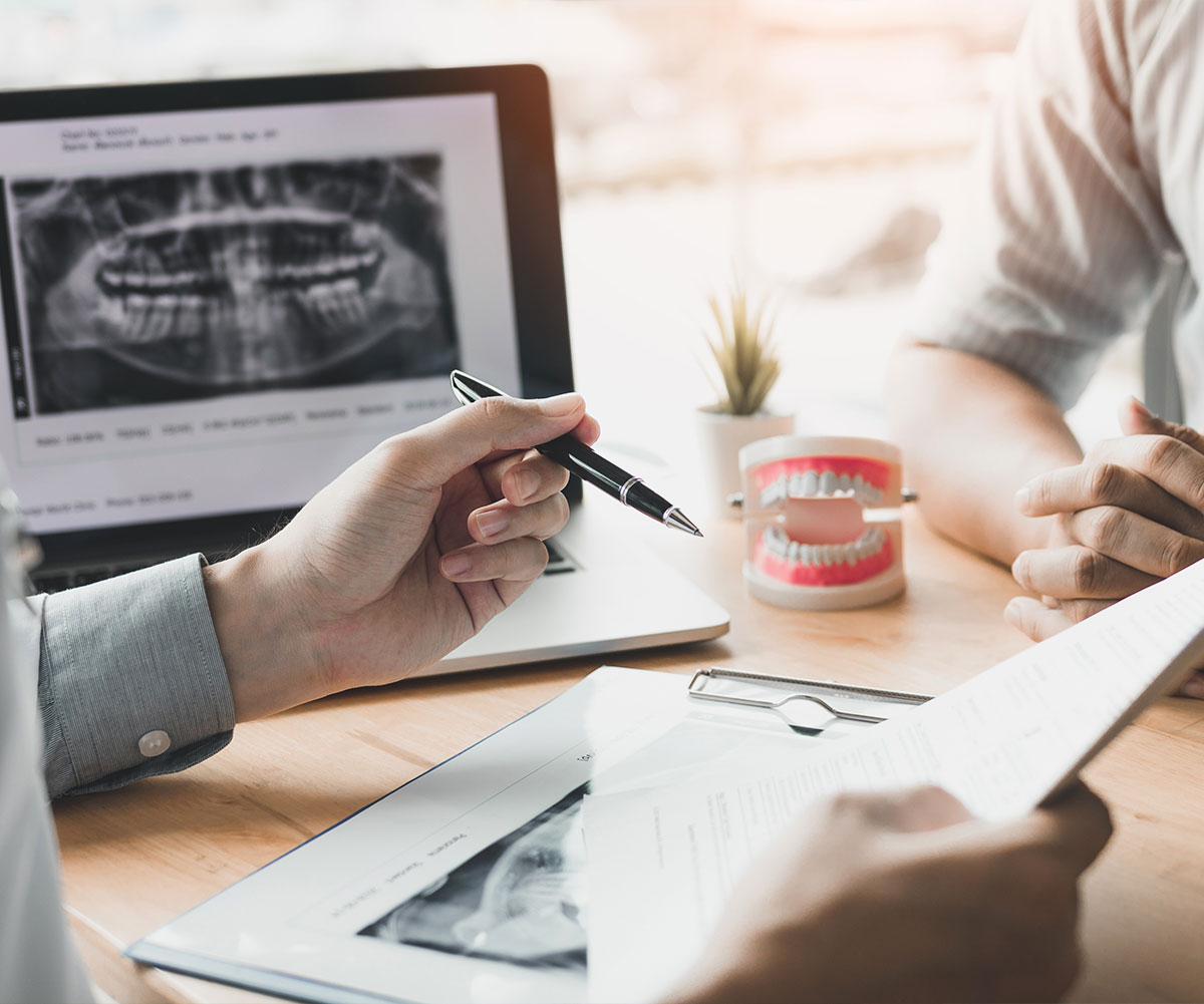 The image shows a professional setting with two individuals reviewing dental imagery on a computer screen, with one person pointing at the image while another writes notes, both seated around a table with a laptop, papers, and a cup.