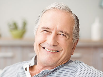 An elderly man with white hair is smiling broadly while sitting comfortably in his home, wearing a blue shirt and a striped tie.